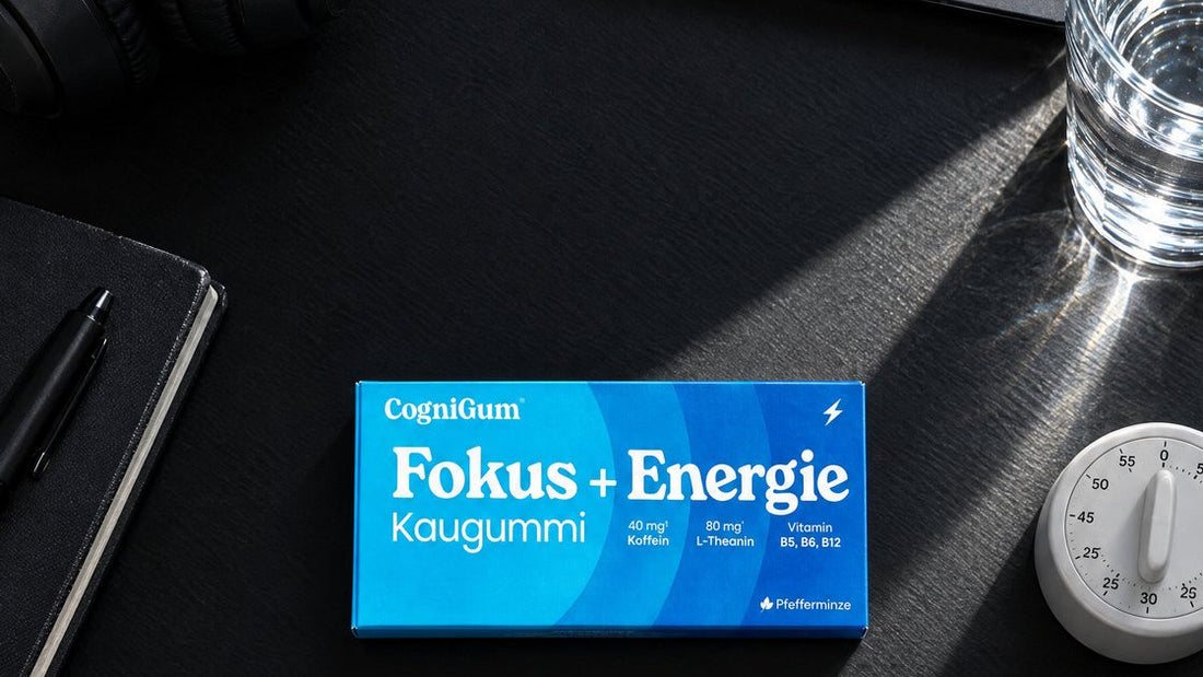 Die CogniGum-Schachtel auf einem schwarzen Schreibtisch zwischen Kopfhörern, Notizbuch mit Stift, einem Glas Wasser und einem Küchenwecker.